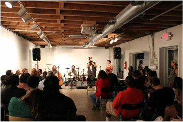 Photo of an open mic. Image is taken from the back of a room and includes the backs of people sitting facing a stage where a group of musicians are performing.
